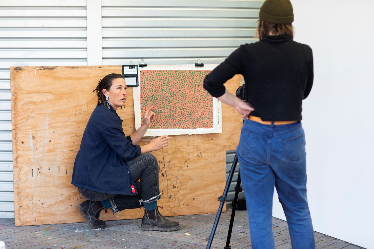 A woman crouching in front of a painting, demonstrating at it, while another woman listens from behind a camera